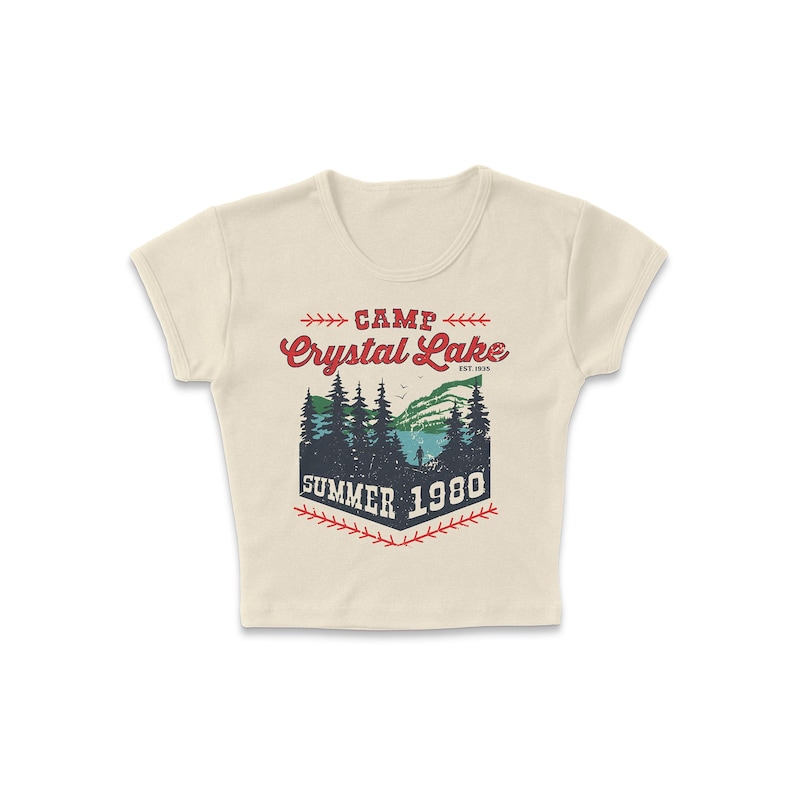 May include: Cream-colored crop top with a graphic design featuring the words "Camp Crystal Lake Summer 1980." The design includes a lake scene with trees and a mountain range, in red, green, and blue.