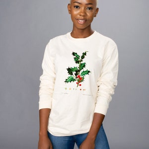 Peut inclure: T-shirt cr&egrave;me &agrave; manches longues avec une illustration botanique de houx, avec des feuilles vertes et des baies rouges. Le t-shirt est port&eacute; avec un jean bleu. L'illustration comprend du texte.