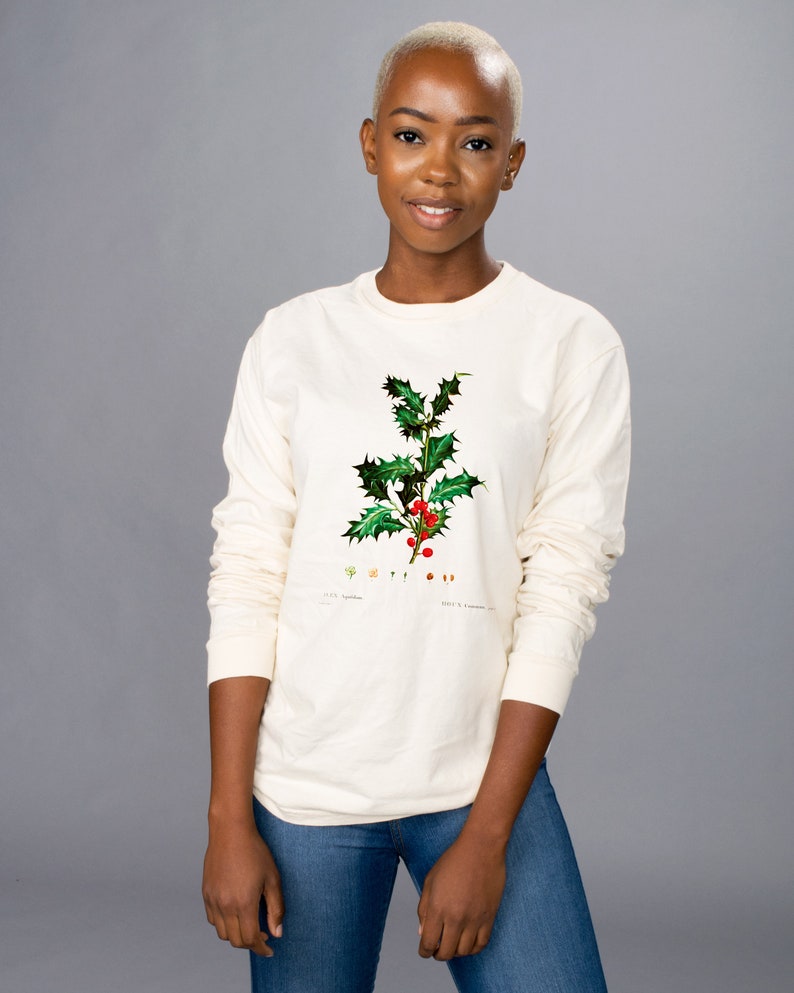 Peut inclure: T-shirt cr&egrave;me &agrave; manches longues avec une illustration botanique de houx, avec des feuilles vertes et des baies rouges. Le t-shirt est port&eacute; avec un jean bleu. L'illustration comprend du texte.