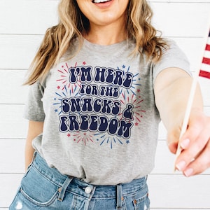Puede incluir: Una camiseta gris con un gráfico azul y rojo que dice "Estoy aquí por las meriendas y la libertad". El gráfico es de estilo retro con fuegos artificiales alrededor del texto.