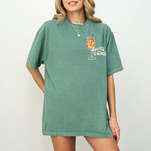 May include: A green short-sleeve t-shirt with a white collar. The shirt features a graphic of an orange cocktail in a glass with the text "Spritz o'Clock" in a retro font. The shirt is a relaxed fit.