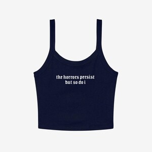 May include: Navy blue crop top with thin straps. The white text on the front reads "the horrors persist but so do i". The top has a scoop neckline and is made of a soft material.
