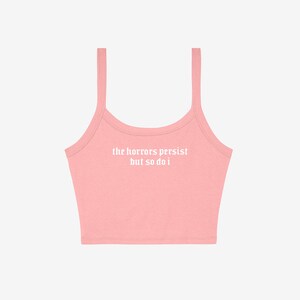 May include: A light pink crop top with thin straps. The text "the horrors persist but so do i" is printed in white across the front. The top is made of a soft, likely cotton, material.