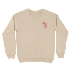 May include: A beige sweatshirt with a pink cherub graphic on the front. The cherub is holding a bow and arrow.