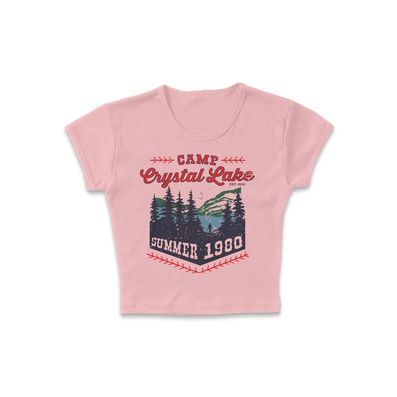 May include: A light pink crop top with a graphic design. The design features the words "Camp Crystal Lake Summer 1980" in red and navy blue, with a scenic illustration of a lake and trees.