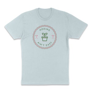 May include: Light blue t-shirt with a green and pink graphic of a plant in a pot with the text "HOEING AIN'T EASY" in a circle.