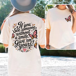 May include: A cream-colored t-shirt featuring a black graphic on the back with the text "If you see someone without a smile, give 'em yours!" surrounded by pink and orange butterflies. A smaller matching butterfly graphic is on the front chest.