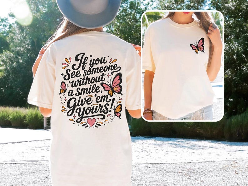 May include: A cream-colored t-shirt featuring a black graphic on the back with the text "If you see someone without a smile, give 'em yours!" surrounded by pink and orange butterflies. A smaller matching butterfly graphic is on the front chest.