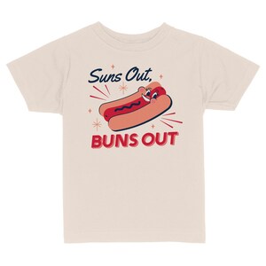 K&ouml;nnte beinhalten: Ein hellbeiges T-Shirt mit einem Cartoon-Hotdog mit einem l&auml;chelnden Gesicht und dem Text "Suns Out, Buns Out" in Rot und Blau.