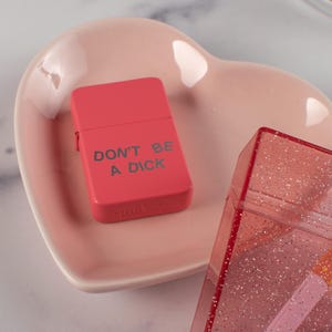 May include: A bright red lighter with the text "DON'T BE A DICK" printed on the front. The lighter is resting on a pink heart-shaped dish. A clear, glittery case is in the background.
