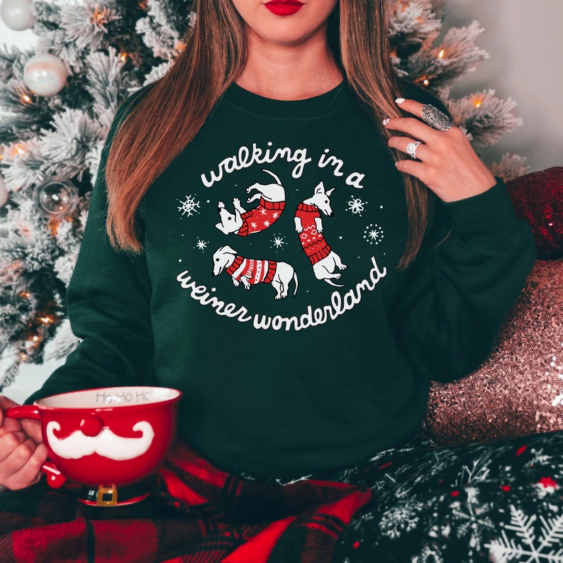 May include: Dark green sweatshirt with the phrase "walking in a wiener wonderland" in white script. The design features four dachshunds wearing red sweaters and snowflakes. A red mug with a white mustache is held in the person's hand.