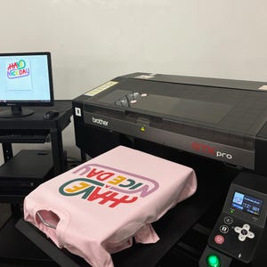 May include: A pink t-shirt with the text "HAVE A NICE DAY" printed in rainbow colours on a Brother GTX Pro direct-to-garment printer.