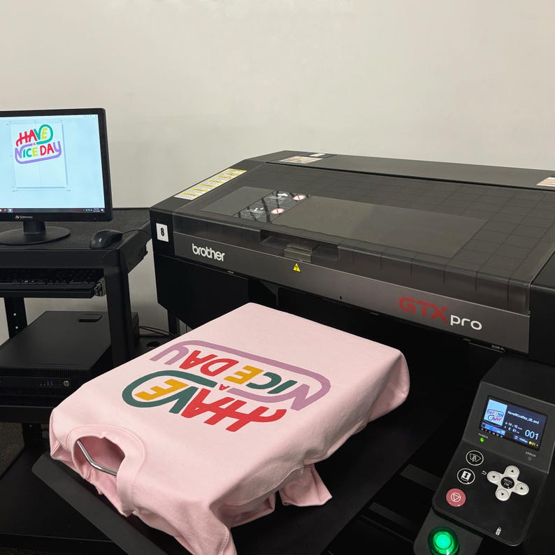 May include: A pink t-shirt with the text "HAVE A NICE DAY" printed in rainbow colours on a Brother GTX Pro direct-to-garment printer.