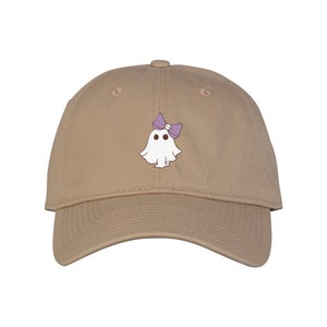 May include: A tan baseball cap featuring a white ghost graphic with a purple bow. The ghost has two black eyes and a small purple bow on its head. The cap is made of a soft material and has a curved brim.