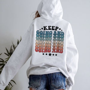 Puede incluir: Sudadera con capucha blanca con un gr&aacute;fico de estilo retro en la espalda que dice "KEEP GOING AND" repetido en un arco&iacute;ris de colores.
