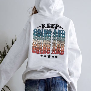 Puede incluir: Sudadera con capucha blanca con un estampado gr&aacute;fico en la espalda que dice "KEEP GOING AND" repetido en un esquema de colores arco&iacute;ris con estrellas en la parte inferior.