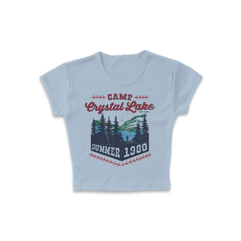 May include: Light blue crop top with a graphic featuring the text "CAMP Crystal Lake SUMMER 1980". The graphic depicts a lake scene with trees and a person. The text is in red and navy blue.