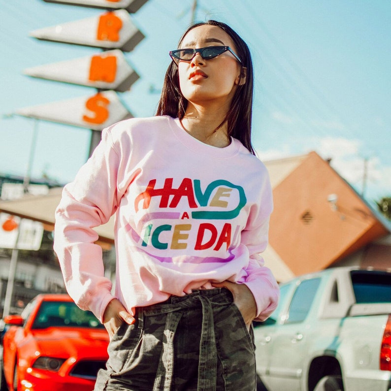 May include: A woman wearing a pink sweatshirt with the text "HAVE A NICE DAY" in colourful letters. She is wearing black sunglasses and has long dark hair. She is standing in front of a red and white sign and a silver car.
