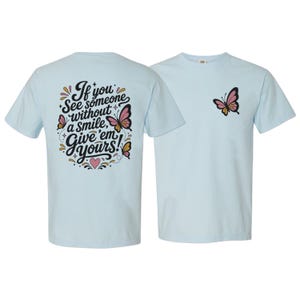 May include: A light blue t-shirt featuring a graphic on the back and front. The back displays the text "If you see someone without a smile, give 'em yours!" surrounded by pink and orange butterflies, stars, and a heart. The front features a single butterfly.