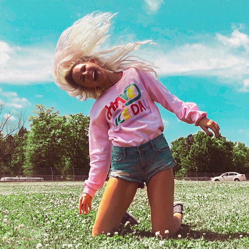 May include: A woman wearing a pink long-sleeved sweatshirt with the text "Have a Nice Day" in rainbow colours. She is wearing denim shorts and has her blonde hair flipped up in the air. She is kneeling in a field of white clover.