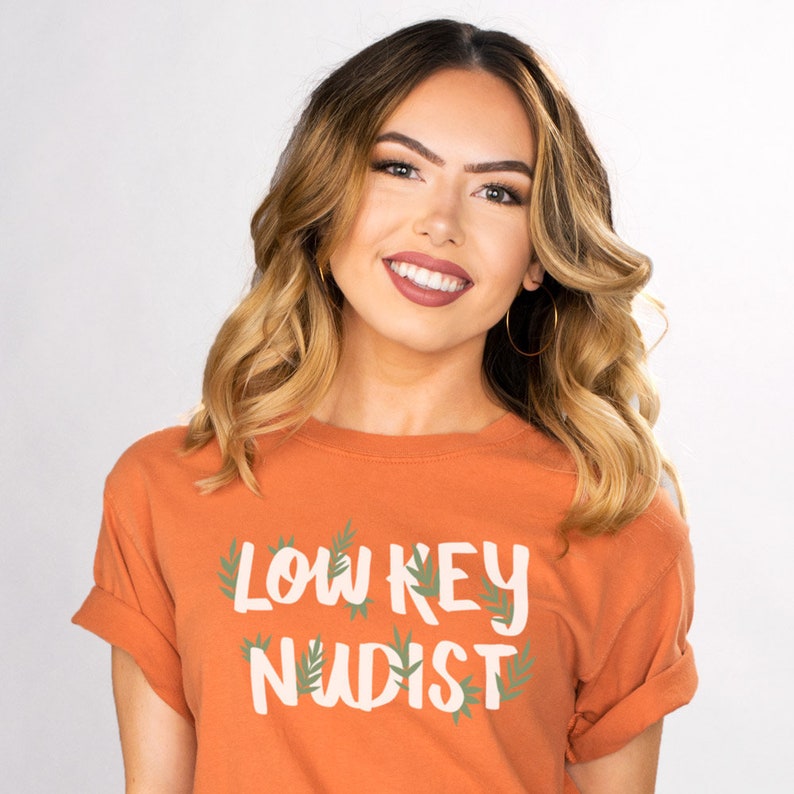 May include: An orange t-shirt with the text "LOW KEY NUDIST" in white, with green leaf accents. The shirt is worn by a person with wavy blonde hair and gold hoop earrings.