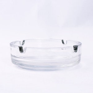 May include: A clear glass ashtray with a round shape and three indentations for cigarettes. The ashtray has a slightly raised lip around the edge.