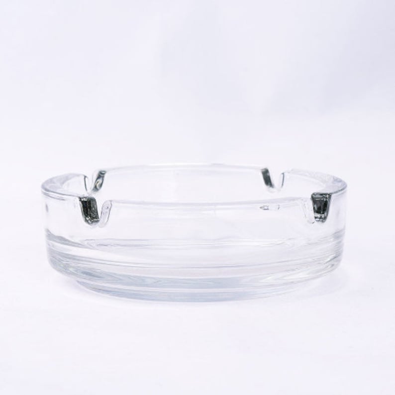 May include: A clear glass ashtray with a round shape and three indentations for cigarettes. The ashtray has a slightly raised lip around the edge.