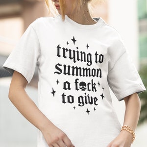 May include: White t-shirt with the phrase "trying to Summon a f*ck to give" in black gothic lettering. The design includes a skull and star accents. The shirt is a casual, oversized style.