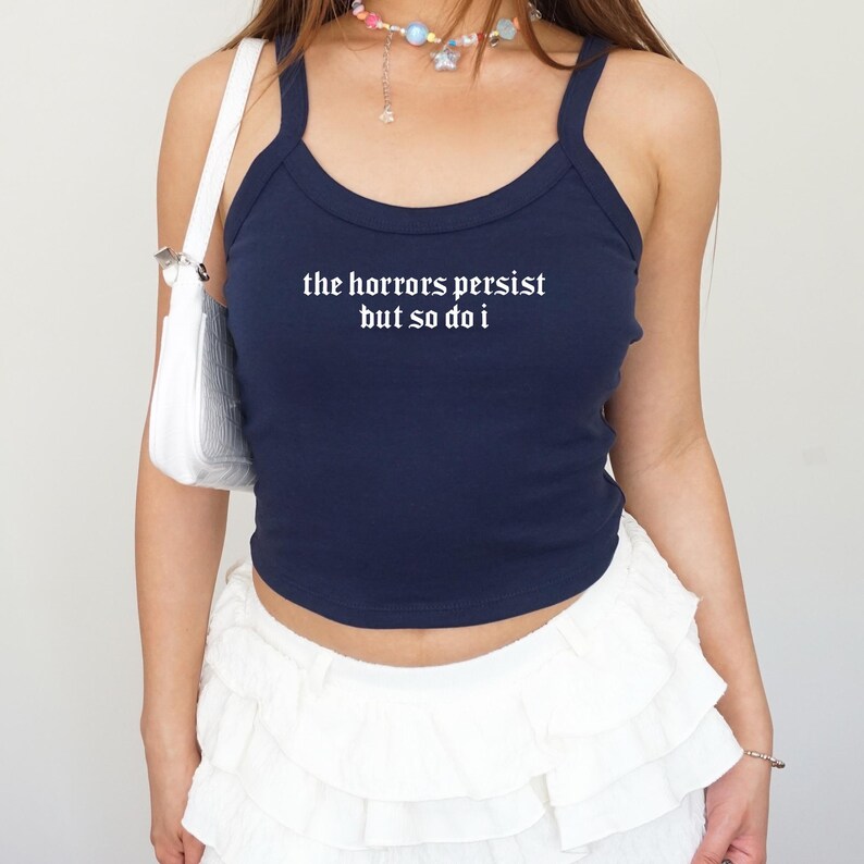 May include: Navy blue tank top with the text "the horrors persist but so do i" in white. The top has thin straps and a scoop neckline. The person is wearing a white ruffled skirt and carrying a white purse.