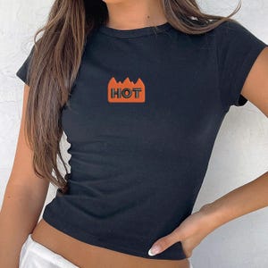 May include: A black short-sleeved t-shirt featuring a central graphic of an orange flame shape with the word HOT printed in bold black capital letters. The garment has a crew neckline and a fitted silhouette, providing a simple and clean design.