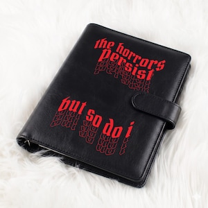May include: Black faux leather planner with the red text "the horrors persist" and "but so do i". The planner has a magnetic closure and is lying on a white, fluffy surface.