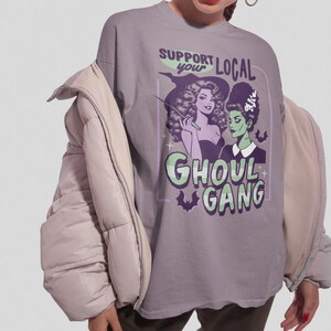 May include: A gray t-shirt featuring a graphic of two women with a witch and monster theme. The text reads "Support Your Local Ghoul Gang" in green and purple. The shirt is a casual style.