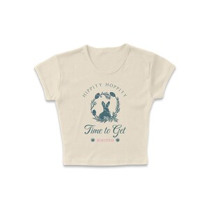 May include: A cream-colored cropped t-shirt with a graphic of a rabbit in a wreath with the text "Hippity Hoppity Time to Get Zooted".
