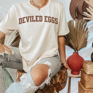 May include: Cream-colored t-shirt with the text "DEVILED EGGS" in brown block letters. The shirt is worn by a person wearing ripped blue jeans. The background includes a red vase, dried plants, and woven baskets.