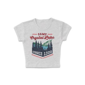 May include: A light gray crop top with a graphic design. The design features the words "CAMP Crystal Lake SUMMER 1980" in red and blue, with a forest and lake scene. The top has short sleeves and a crew neck.
