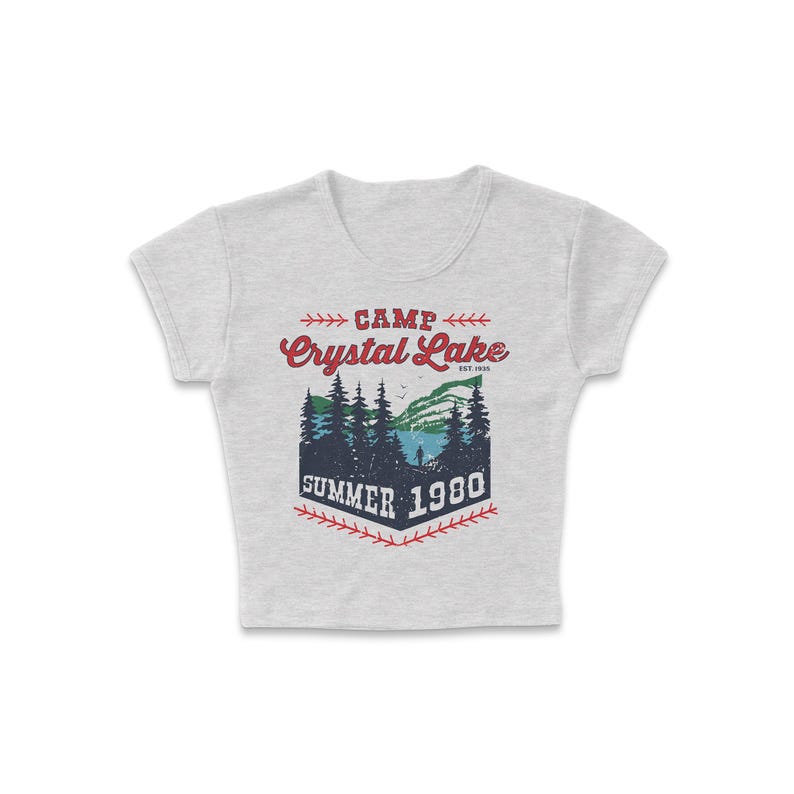 May include: A light gray crop top with a graphic design. The design features the words "CAMP Crystal Lake SUMMER 1980" in red and blue, with a forest and lake scene. The top has short sleeves and a crew neck.