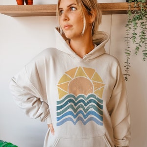 Puede incluir: Una sudadera con capucha de color crema con un gr&aacute;fico de estilo vintage de un sol poni&eacute;ndose sobre agua ondulada. El gr&aacute;fico est&aacute; en tonos de amarillo, naranja, azul y verde.