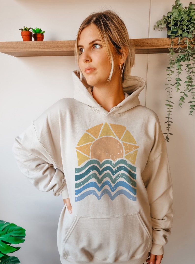 Puede incluir: Una sudadera con capucha de color crema con un gr&aacute;fico de estilo vintage de un sol poni&eacute;ndose sobre agua ondulada. El gr&aacute;fico est&aacute; en tonos de amarillo, naranja, azul y verde.