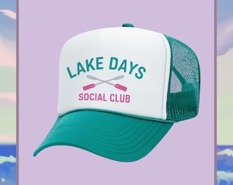 May include: A teal and white trucker hat with the text "LAKE DAYS SOCIAL CLUB" in teal and pink. The hat features a teal brim and mesh back, with a white front panel. The design includes crossed oars.