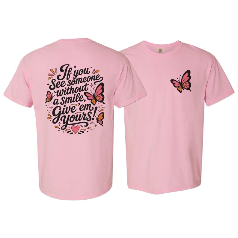 May include: A light pink t-shirt featuring a small butterfly graphic on the front left chest. The back displays a large black script text that reads, If you see someone without a smile, Give 'em yours!, surrounded by butterflies and hearts.