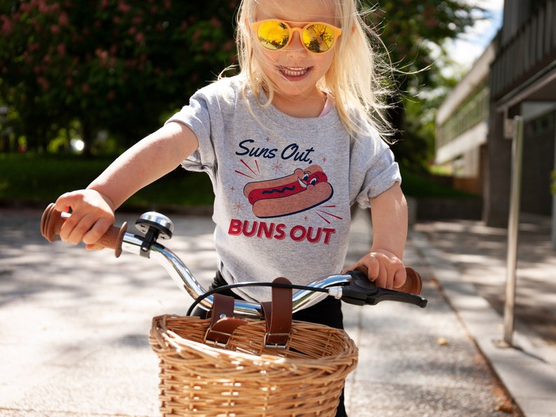 K&ouml;nnte beinhalten: Ein junges Kind tr&auml;gt ein graues T-Shirt mit einem Hotdog-Motiv und dem Text "Suns Out, Buns Out" und f&auml;hrt ein kleines Fahrrad mit einem Weidenkorb.