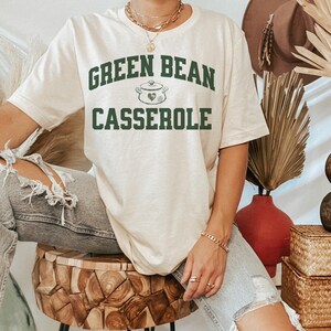 May include: Cream-colored t-shirt with the text "GREEN BEAN CASSEROLE" in dark green, along with a pot graphic. The shirt is a casual style, suitable for everyday wear. The design is centered on the front of the shirt.