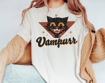 May include: Cream-colored t-shirt featuring a vintage-style graphic of a black cat with a vampire-like grin inside a brown triangle. The word "Vampurr" is written in a retro font below the cat. The design includes star accents.
