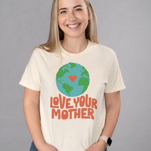 May include: A cream colored t-shirt with a green and blue globe graphic with a red heart in the center. The text "LOVE YOUR MOTHER" is printed in orange below the globe.