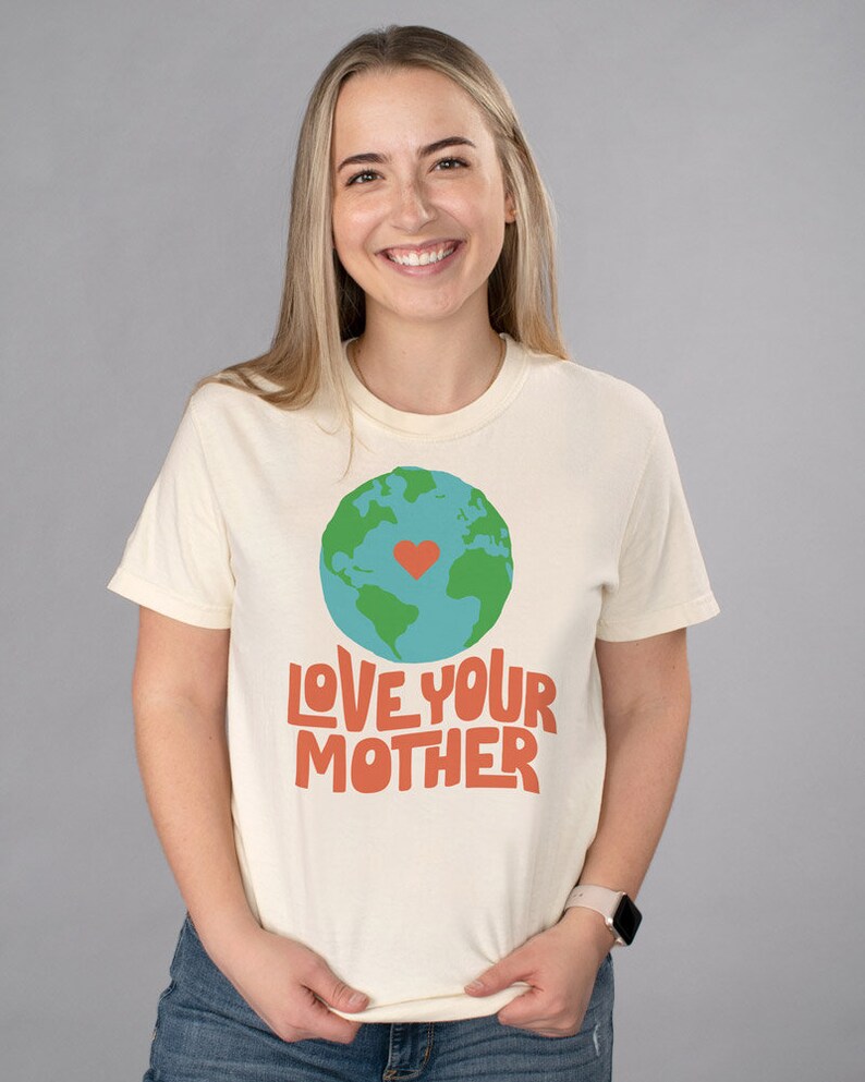 May include: A cream colored t-shirt with a green and blue globe graphic with a red heart in the center. The text "LOVE YOUR MOTHER" is printed in orange below the globe.