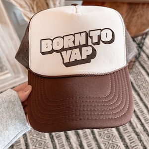 May include: A brown and white trucker hat with a brown mesh back. The front of the hat has the text "BORN TO VAP" in a bold, white font.