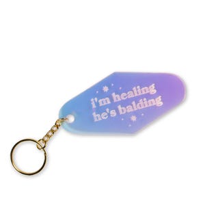 May include: A motel-style keychain with a gradient blue and purple background. The keychain has a gold chain and ring. The text "i'm healing he's balding" is printed in white.