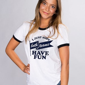 Puede incluir: Camiseta blanca con mangas negras de ribete, con un gr&aacute;fico azul que dice "I Just Hope Both Teams Have Fun" con un dise&ntilde;o de bandera vintage.