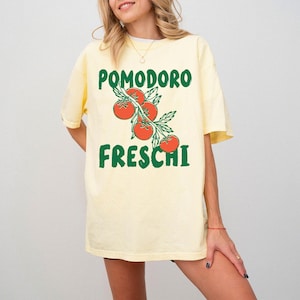 May include: A light yellow t-shirt with the words "POMODORO FRESCHI" in green, along with a graphic of red tomatoes on a vine. The shirt is a casual, oversized fit.