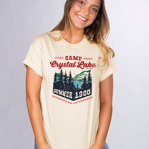 May include: A light yellow t-shirt with a vintage-style graphic design. The design features a forest scene with a lake, trees, and a person fishing. The text reads "Camp Crystal Lake Est. 1925 Summer 1980".